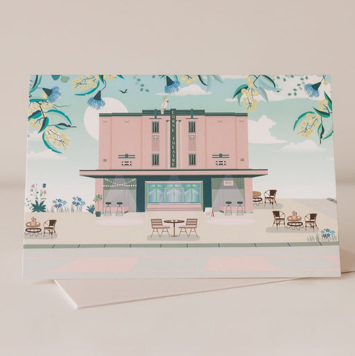 A greeting card with a illustration of a pink theatre building, with a blue floral pattern in the background. The card is positioned vertically.